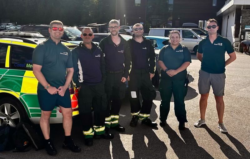 Six people in uniform stand outdoors in a parking lot next to an ambulance vehicle, posing for a group photo.