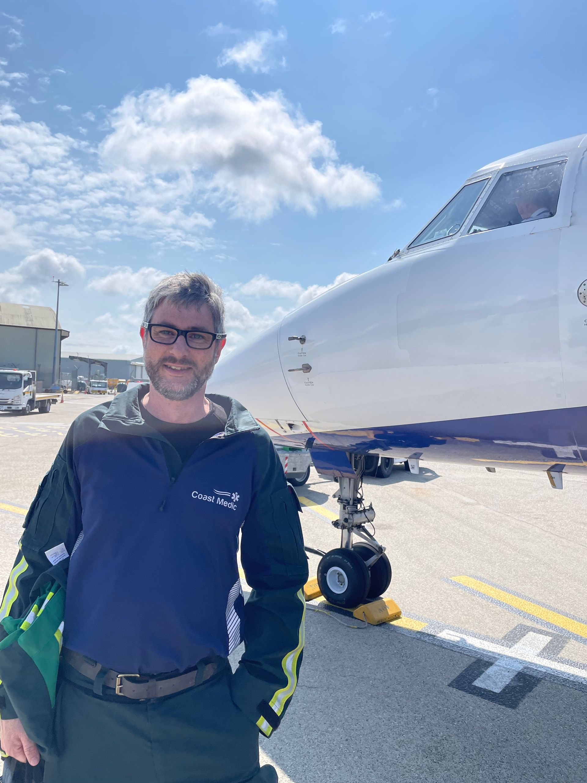 A person wearing a dark jacket stands smiling on an airport tarmac next to the nose and landing gear of a parked plane.