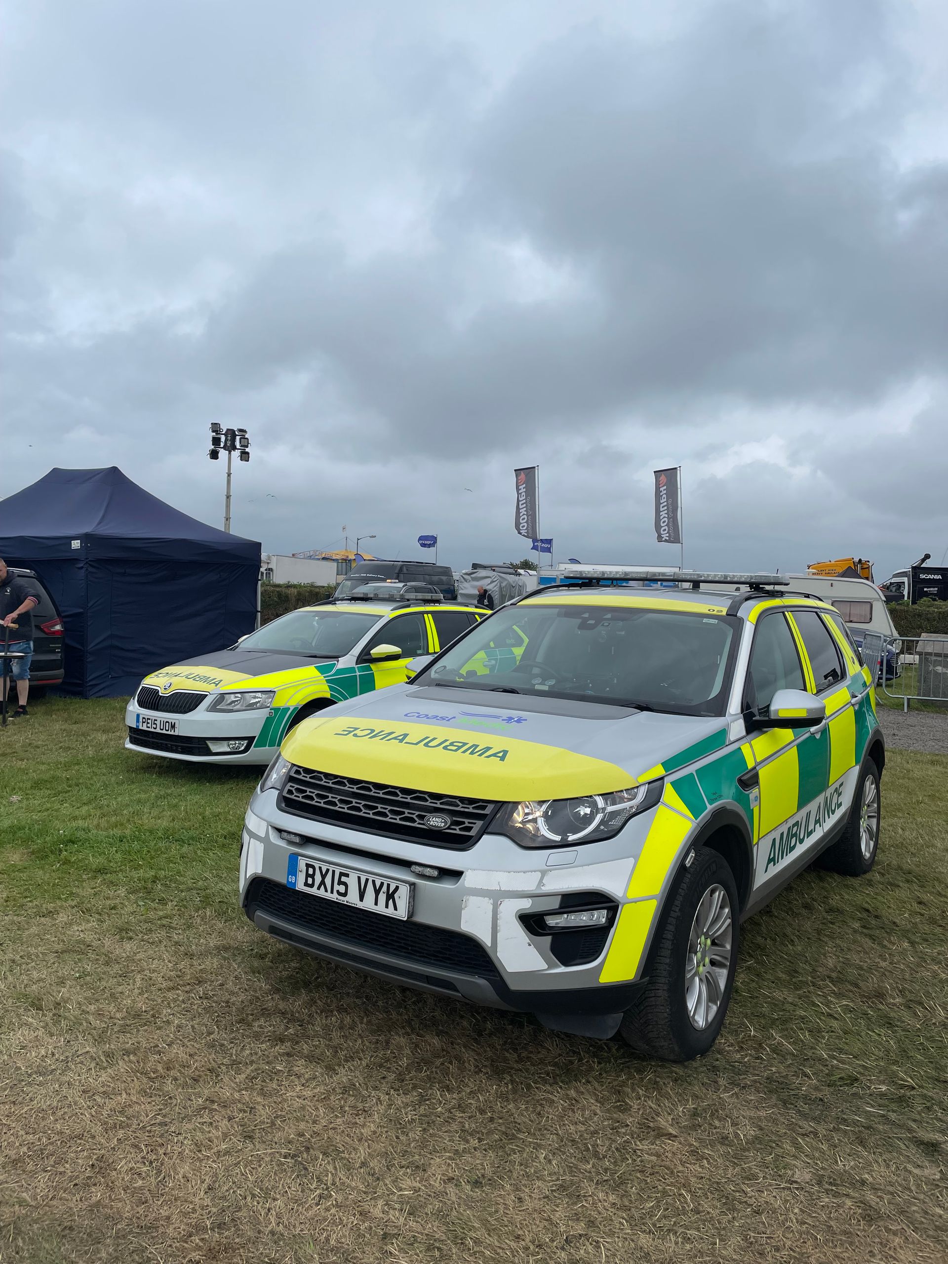 Two emergency vehicles with yellow and green reflective livery parked on a grass field under a cloudy sky.