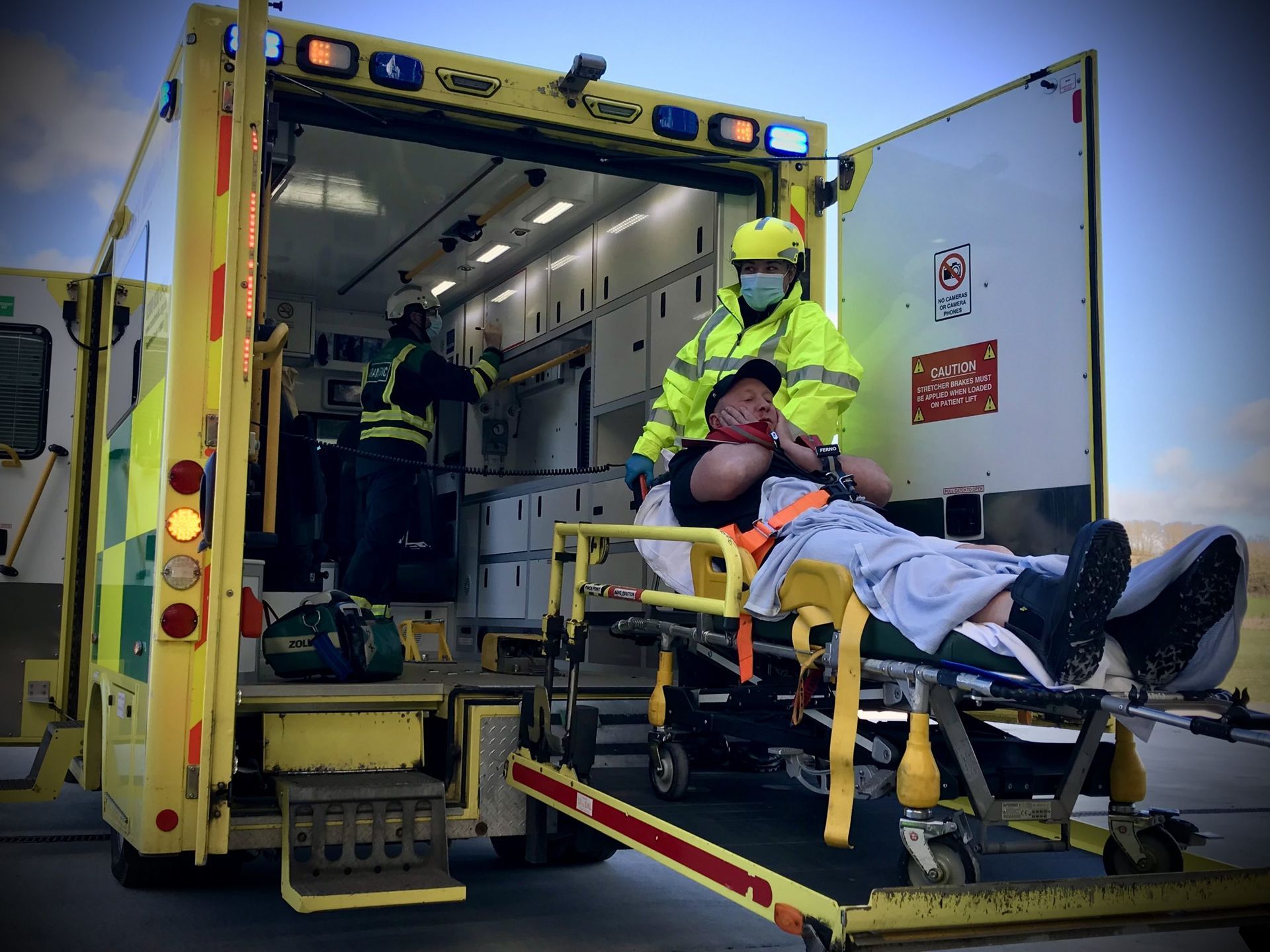 Paramedics wearing high-visibility gear assist a patient on a stretcher into the back of an open ambulance.
