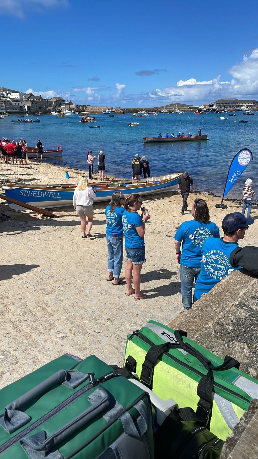 People gather on a rocky beach by the sea, preparing boats for rowing near a town with blue sky above.
