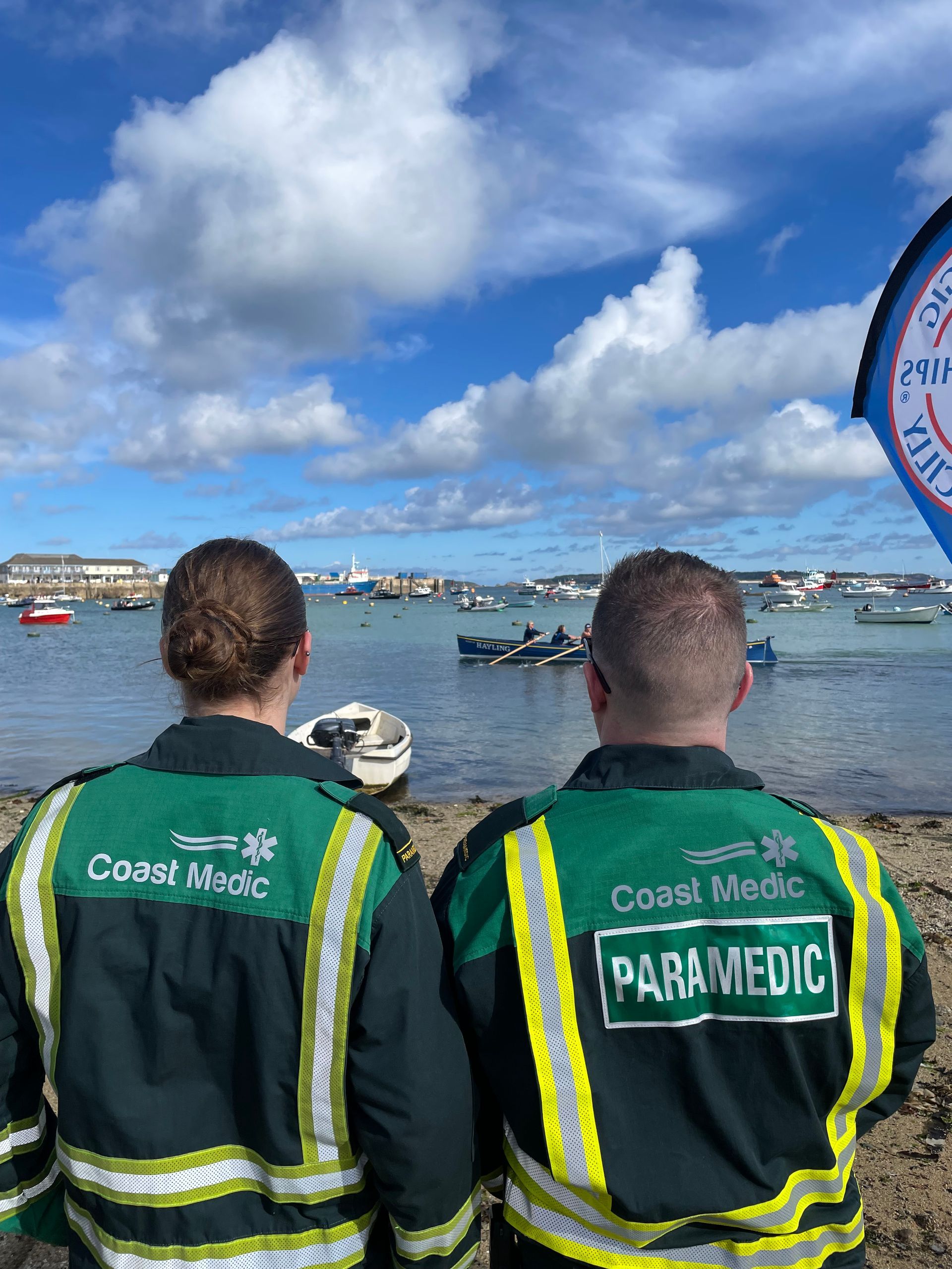 Two people in green Coast Medic jackets look out over a harbor with small boats under a partly cloudy sky.