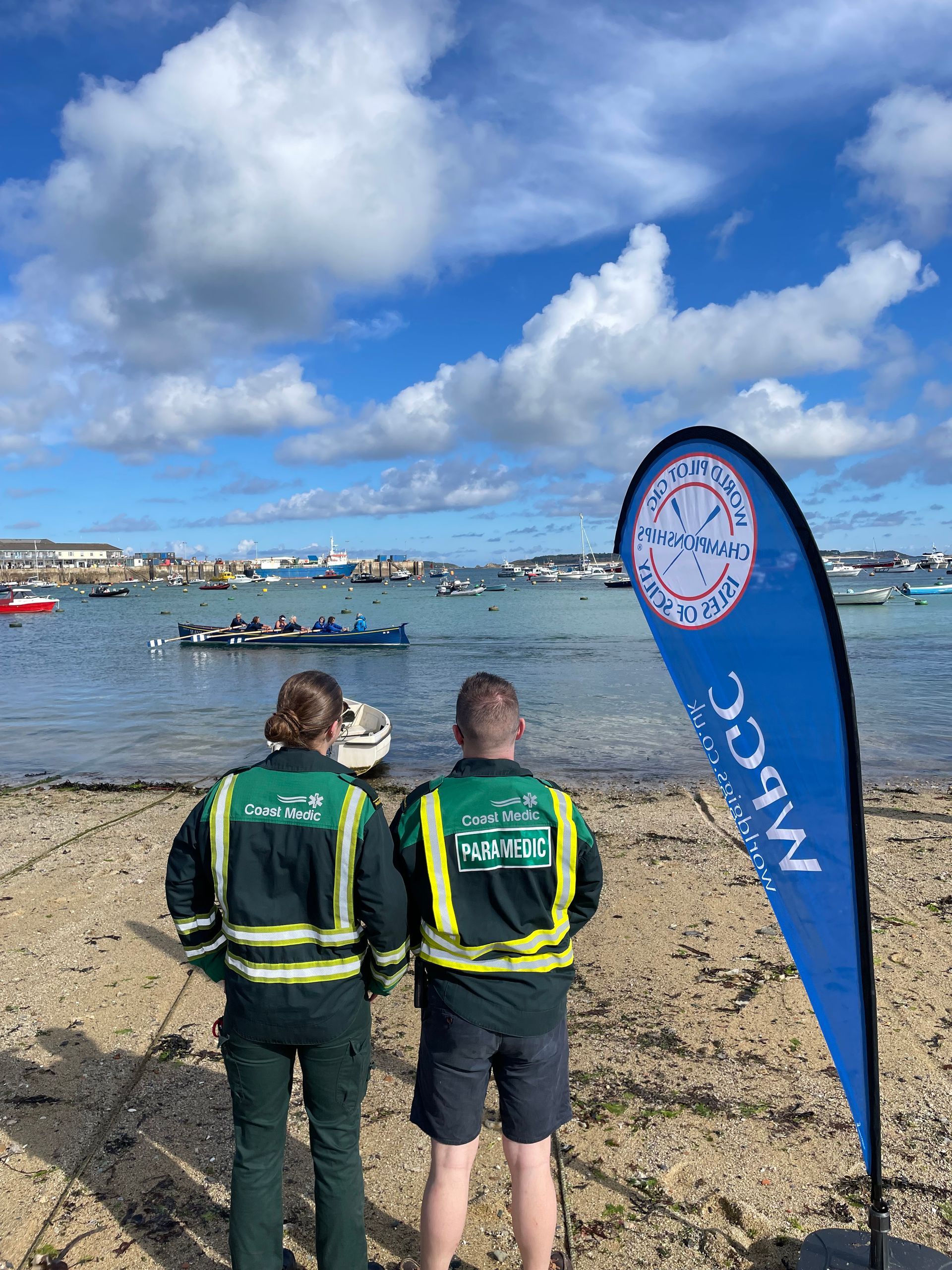 Two people in high-visibility uniforms stand on a rocky beach, looking toward boats in a harbor under a cloudy blue sky.