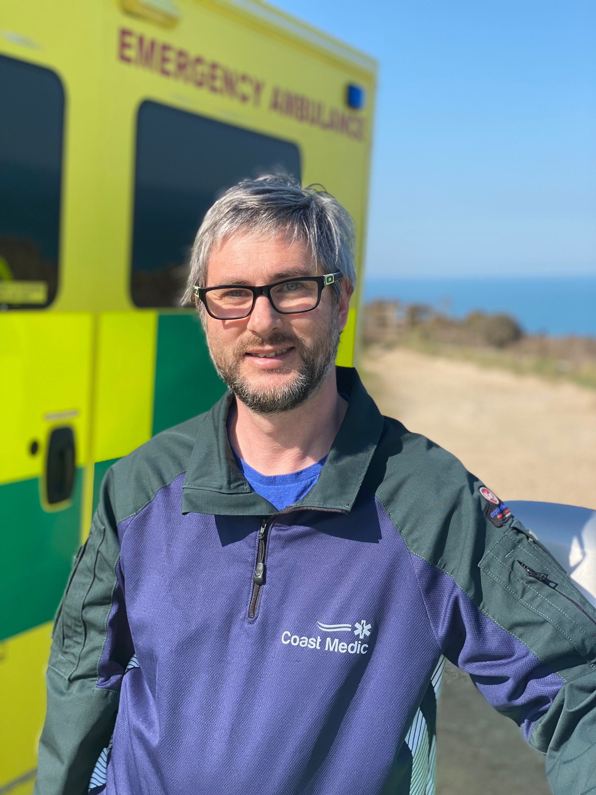 A person in a green and purple uniform stands in front of a yellow emergency ambulance with a coastal view.