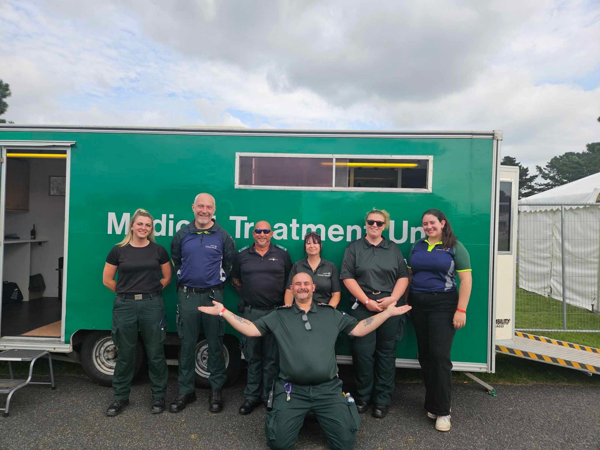 Six people in uniform stand and kneel in front of a green Medical Treatment Unit trailer on a gravel lot.