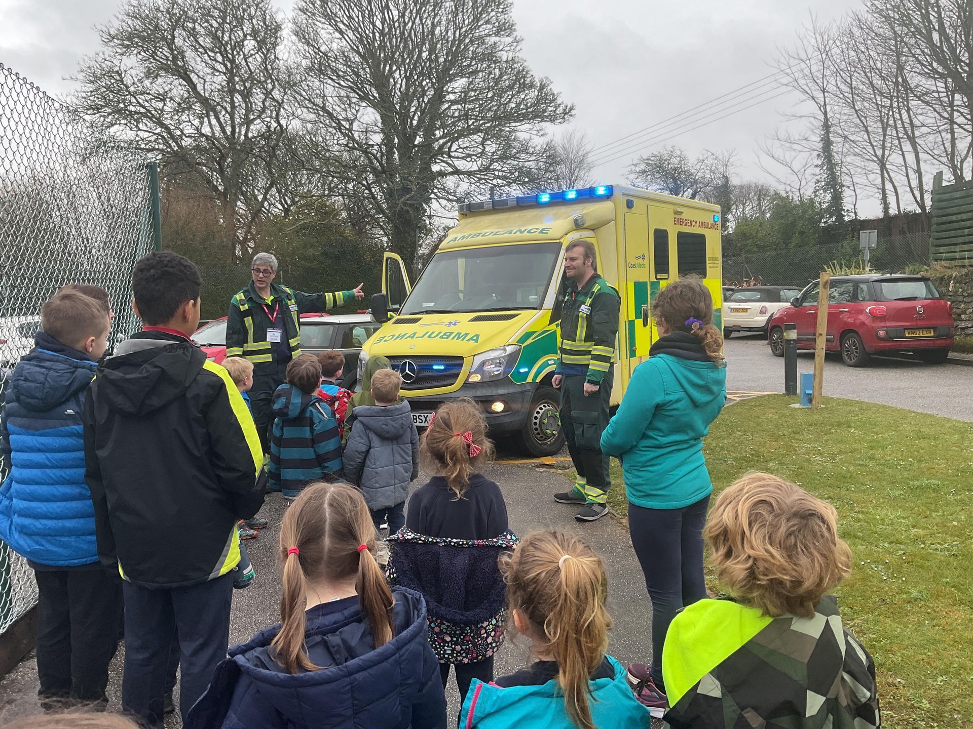 Two emergency medical workers stand by a yellow ambulance, speaking to a group of children outdoors on a cloudy day.