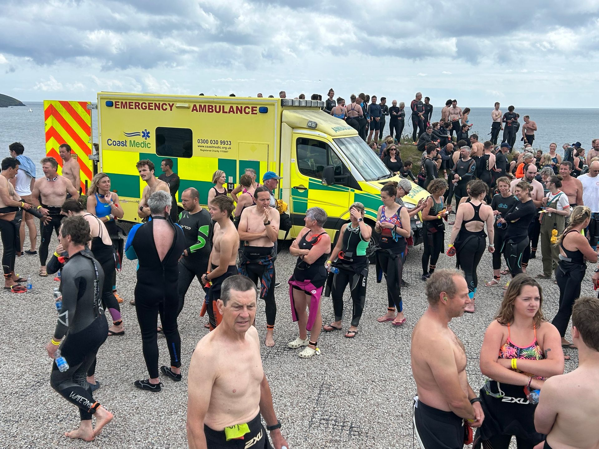 A yellow ambulance stands on a rocky beach near a crowd of people in wetsuits or swimwear at an outdoor swimming event.