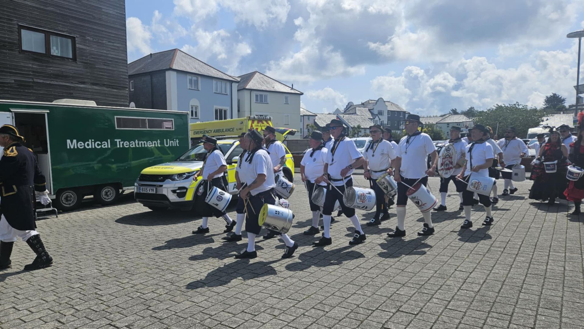 A marching band in white shirts and black pants plays drums on a paved street beside a medical unit vehicle.