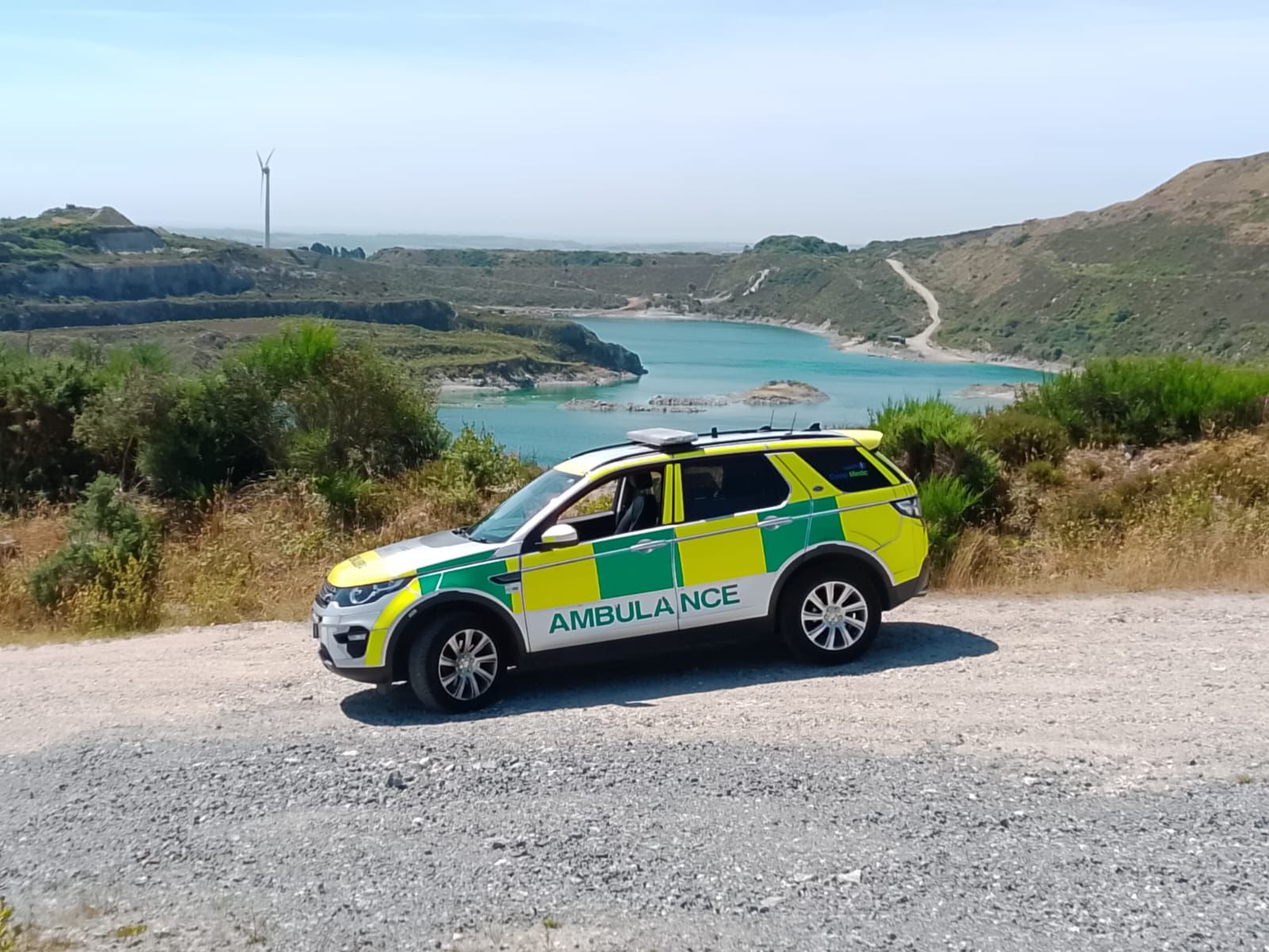 A yellow and white emergency ambulance vehicle parked on a gravel path overlooking a scenic lake and hills.