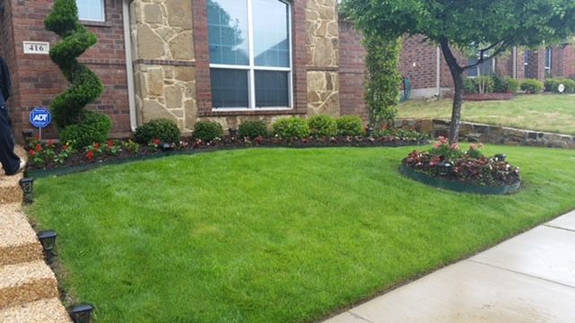 Manicured front lawn with flower beds beside a brick house and wet sidewalk