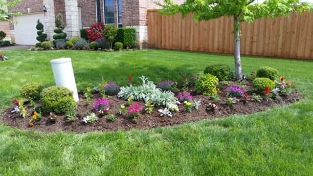 Colorful flower bed with pink, white, and purple blooms in a landscaped front yard beside a lawn and brick house