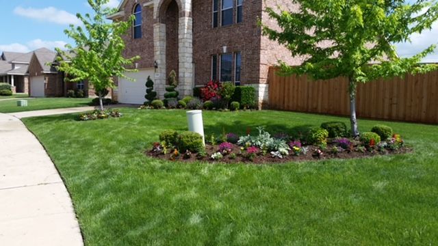 Suburban brick home with a curved sidewalk, green lawn, flower bed, and young trees