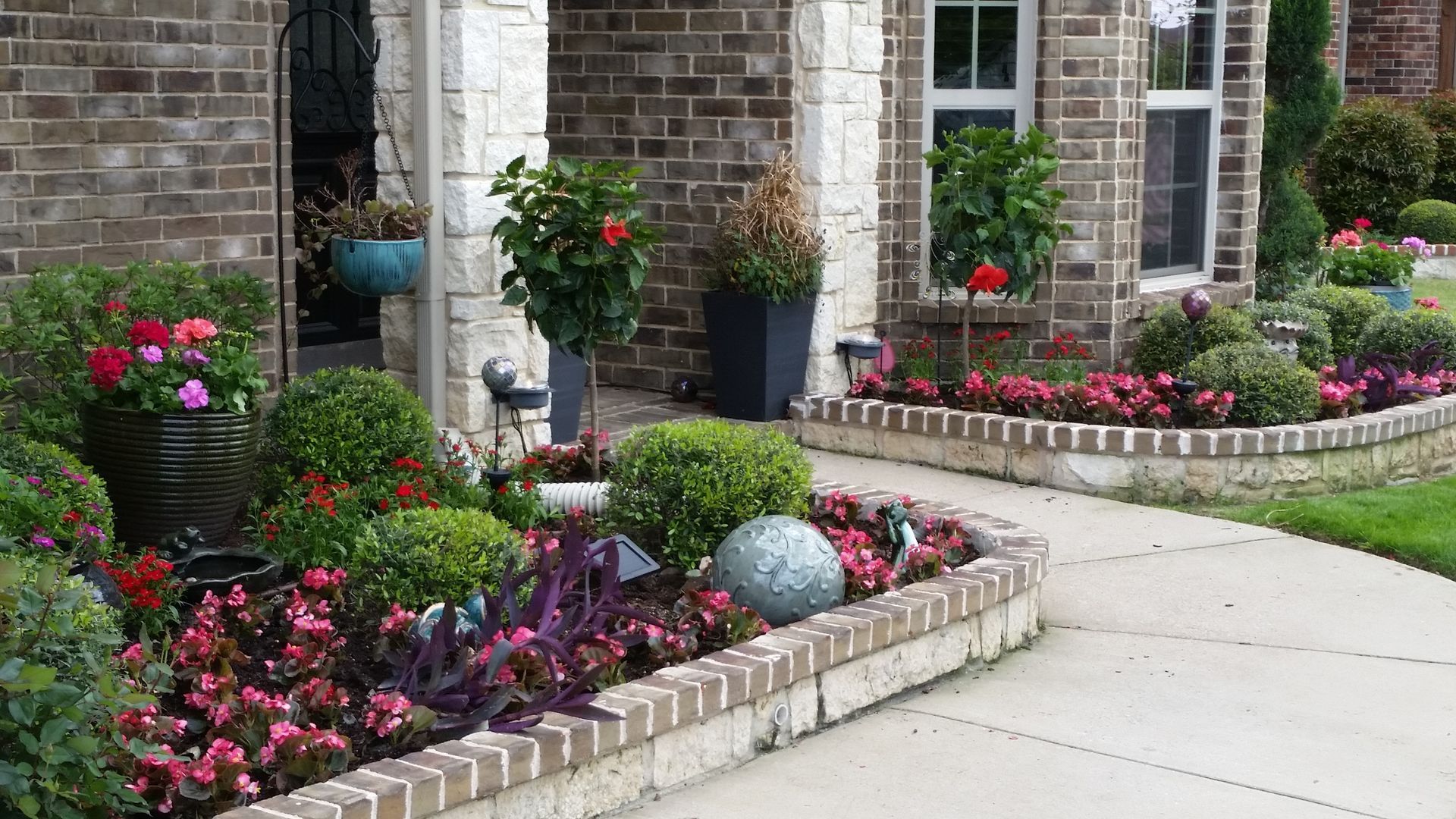 Curved brick flower bed with colorful blooming plants beside a stone house and driveway