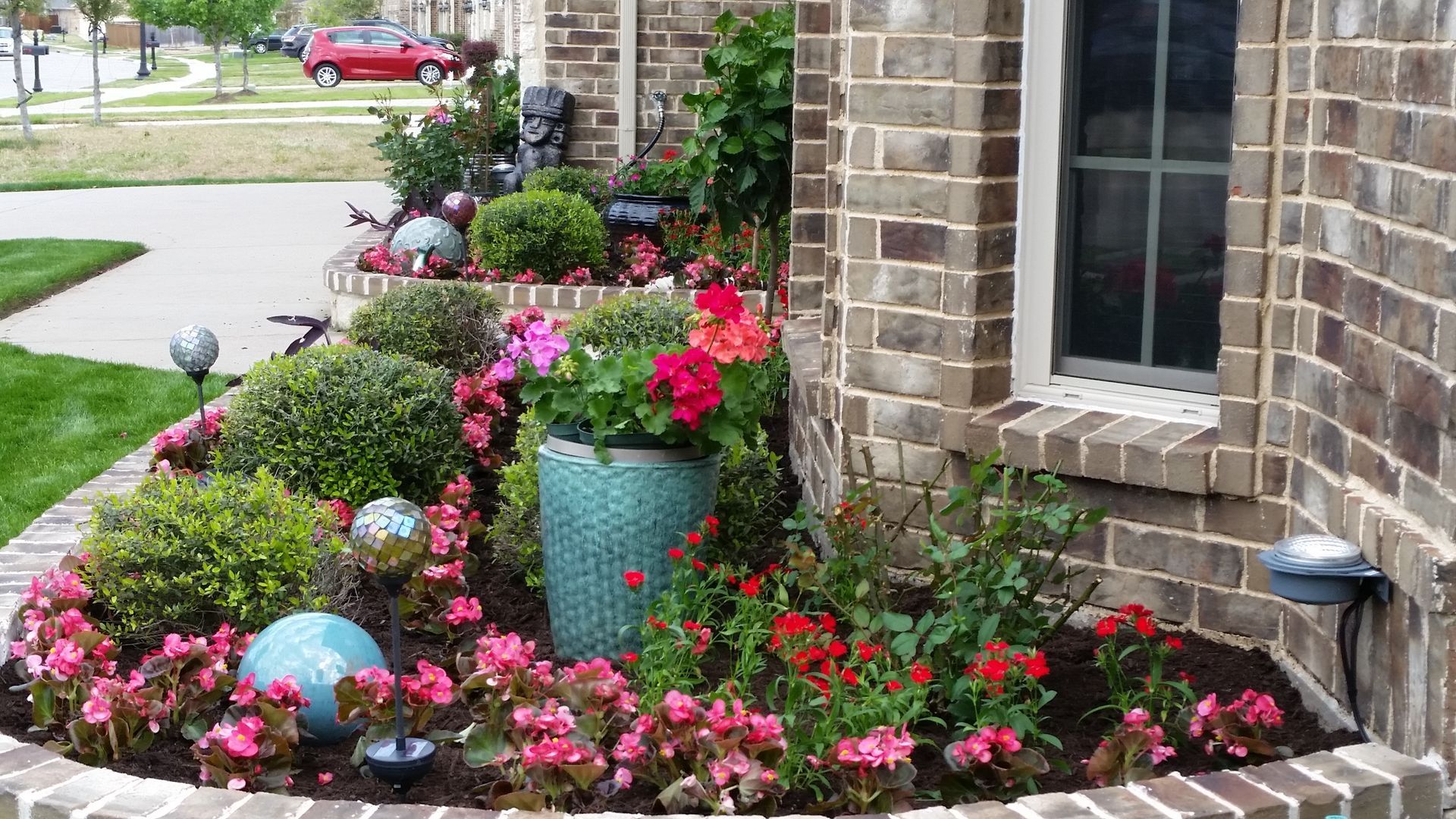 Colorful flower bed with pink blooms and decorative pots beside a brick house and sidewalk