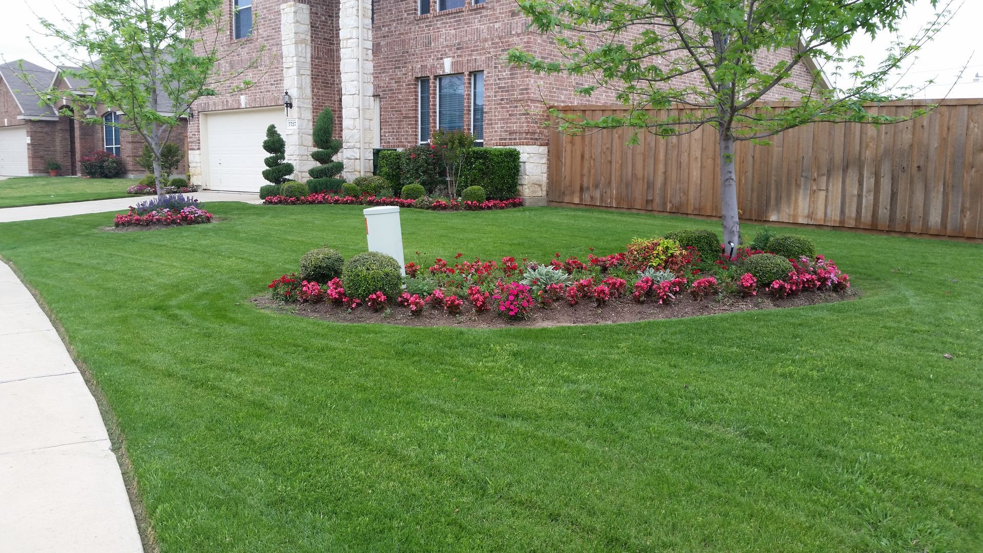 Manicured front yard with green lawn, red flower beds, a mailbox, and a brick house beside a sidewalk