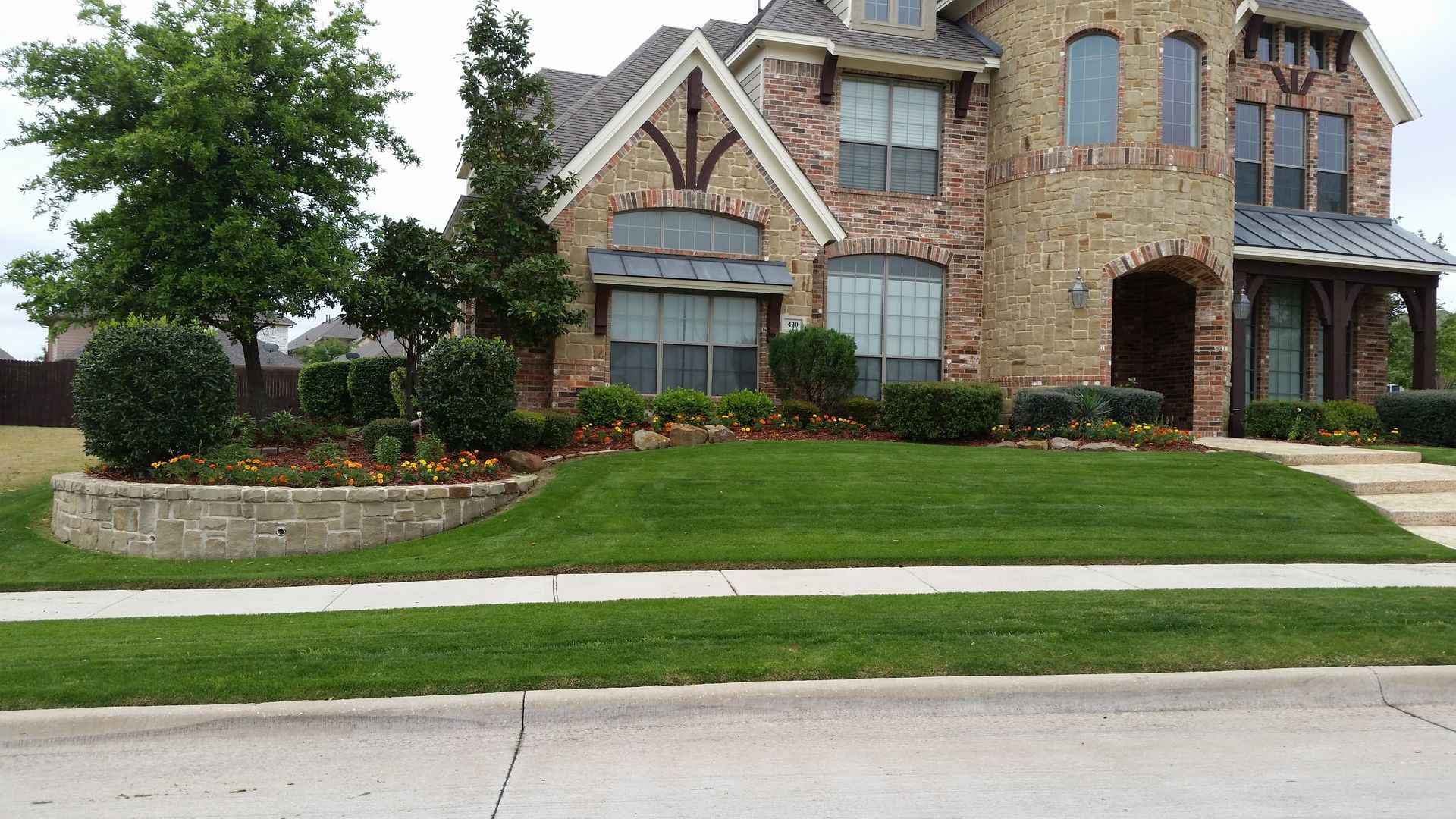 Large stone suburban house with manicured lawn, curved retaining wall, and sidewalk along the street
