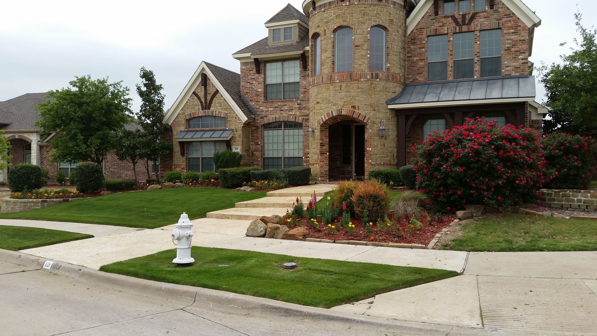 Stone suburban house with front lawn, sidewalk, flower beds, and a white fire hydrant at the curb