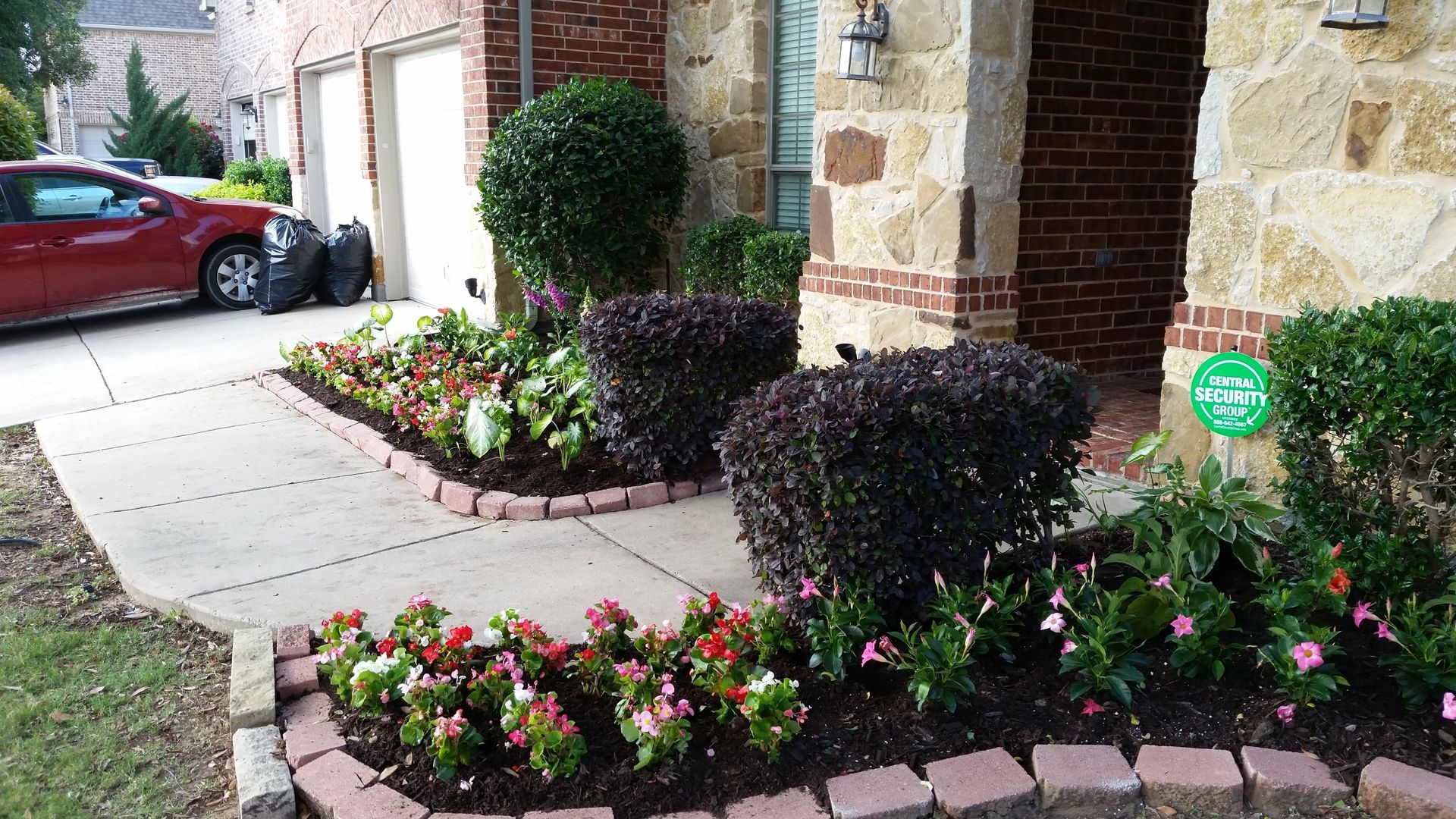 Front yard garden with pink flowers, brick edging, walkway, and a red car parked by the house