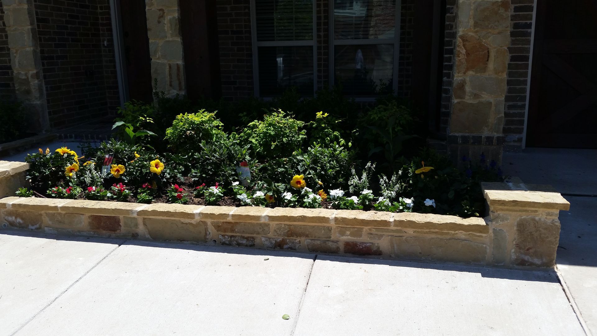 Brick planter with colorful flowers and green shrubs beside a sidewalk in front of a building