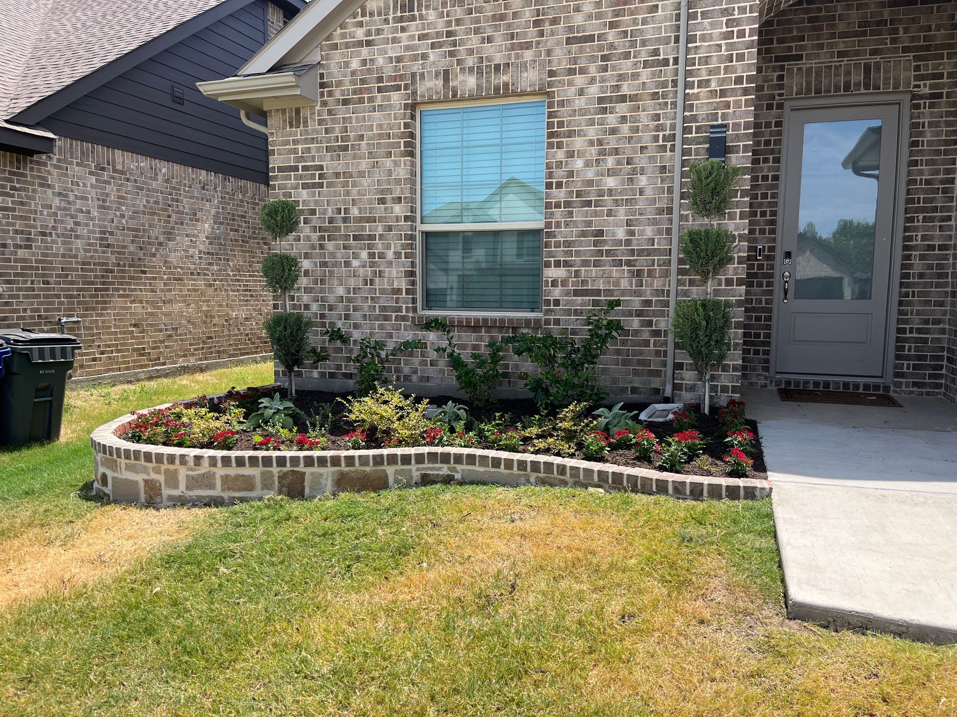 Brick house front yard with curved flower bed, green lawn, and a gray front door at the entrance