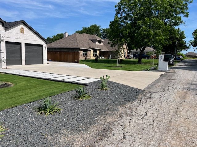Residential driveway and garage beside a gravel lot, with a sidewalk and landscaped front yard on a sunny day