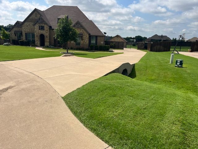 Large brick house with a curved concrete driveway over a drainage culvert in a green suburban yard