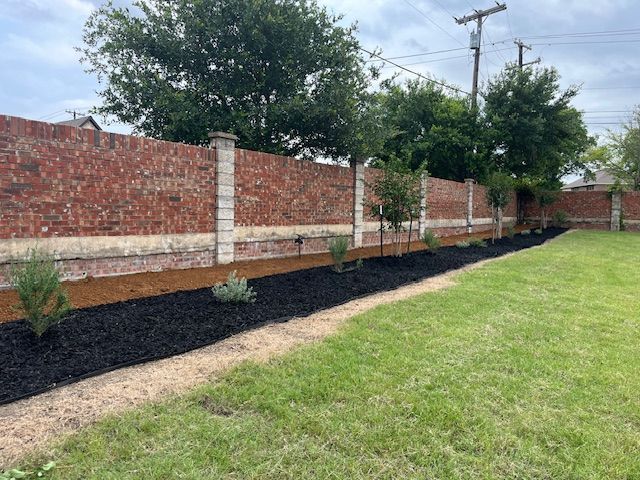 Brick fence beside a green lawn with black mulch and young shrubs under a cloudy sky