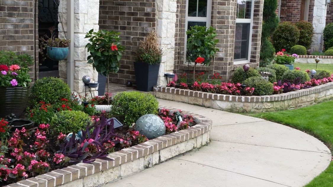 Brick garden border with pink and red flowers along a curved sidewalk outside a stone house