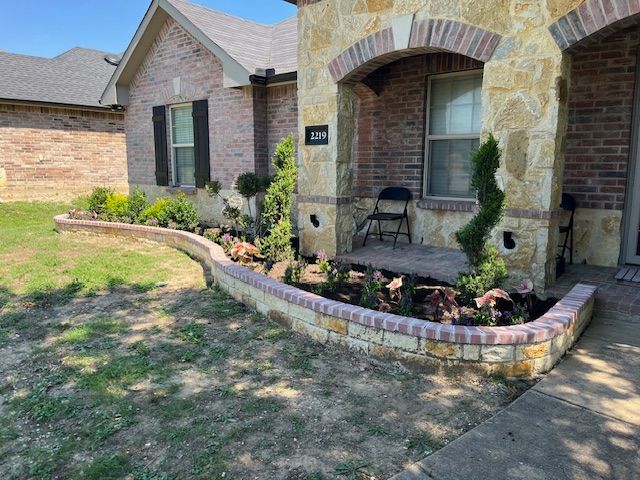Brick house front porch with curved flower bed, black chair, and small shrubs in the yard