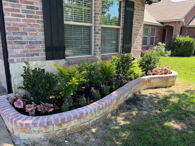 Curved brick flower bed with pink blooms and shrubs beside a brick house and windows