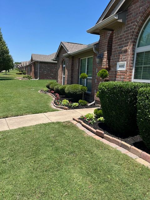 Brick suburban houses with manicured lawns, hedges, and a sidewalk on a sunny day