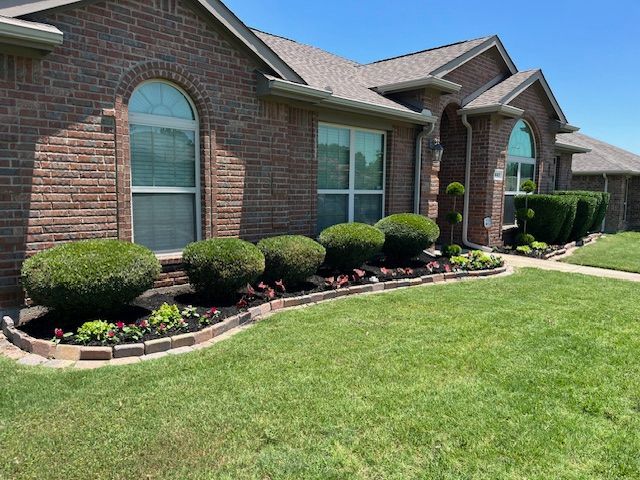 Brick suburban house with arched window, trimmed shrubs, flower beds, and a green lawn under a blue sky