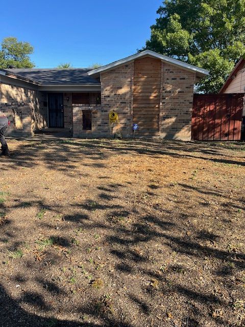 Single-story brick house with a dry front yard, trees, and a red shed in bright sunlight