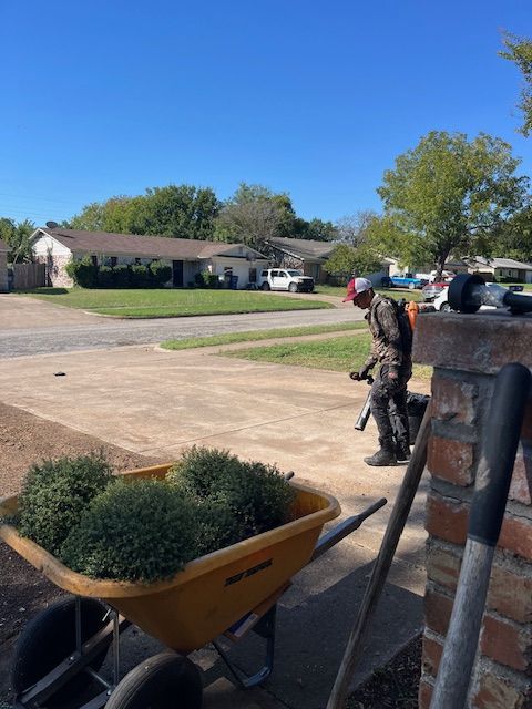 Worker in safety gear on a suburban street beside a wheelbarrow and shovel, near a lawn under a clear blue sky