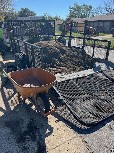Dump trailer loaded with dirt and a wheelbarrow beside it on a sunny residential street.