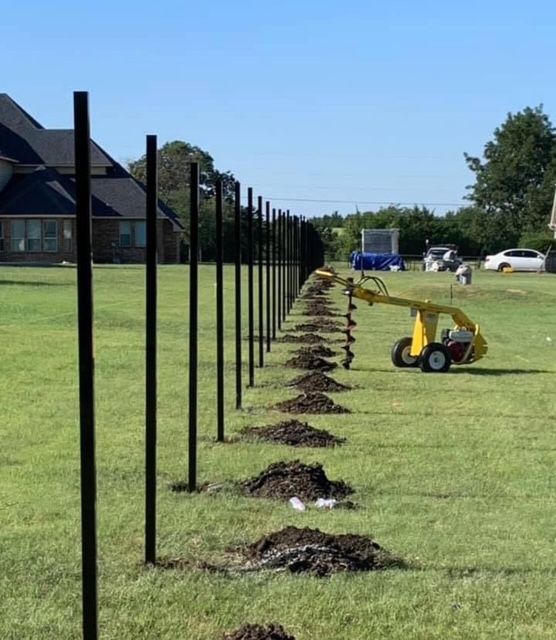 Black fence posts line a grassy field with fresh dirt piles; a small yellow trencher sits nearby.