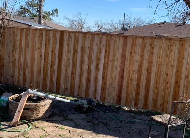 Wooden privacy fence in a backyard with dirt ground, plants, and neighboring roofs visible behind it.