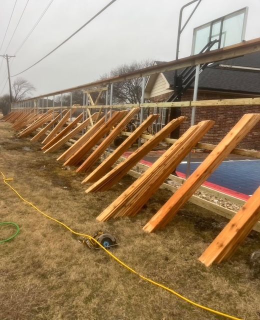 Rows of wooden roof braces leaning against a building at a construction site, with a yellow cord on the ground