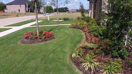 Suburban front yard with green lawn, flower beds, and a sidewalk beside a house