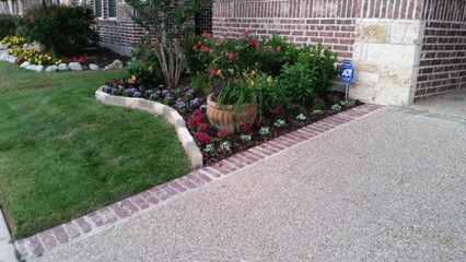 Front yard landscaping with grass, flowers, shrubs, and a brick-edged driveway beside a house.
