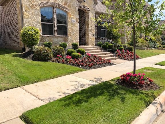 Brick house entrance with stone steps, trimmed lawn, and red flower beds along the sidewalk