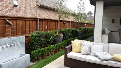 Backyard patio with wicker sofa, water feature, and trimmed hedges beside a wooden fence.