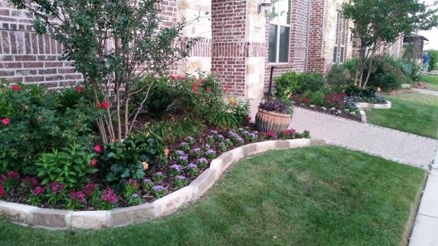 Curved flower bed with pink blooms and shrubs beside a brick house and sidewalk