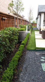 Narrow garden path beside a wooden fence, with green shrubs, gravel, and a white house wall.