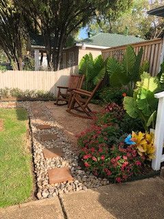 Backyard garden with wooden chairs, brick path, and colorful flowering plants beside a lawn