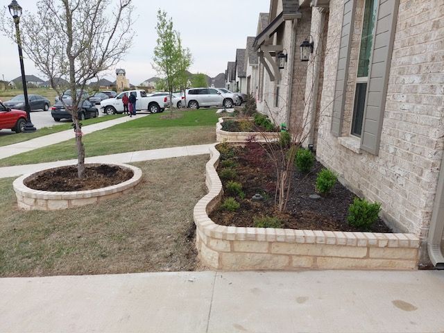 Sidewalk beside a stone building with raised flower beds and a young tree, near a parking lot