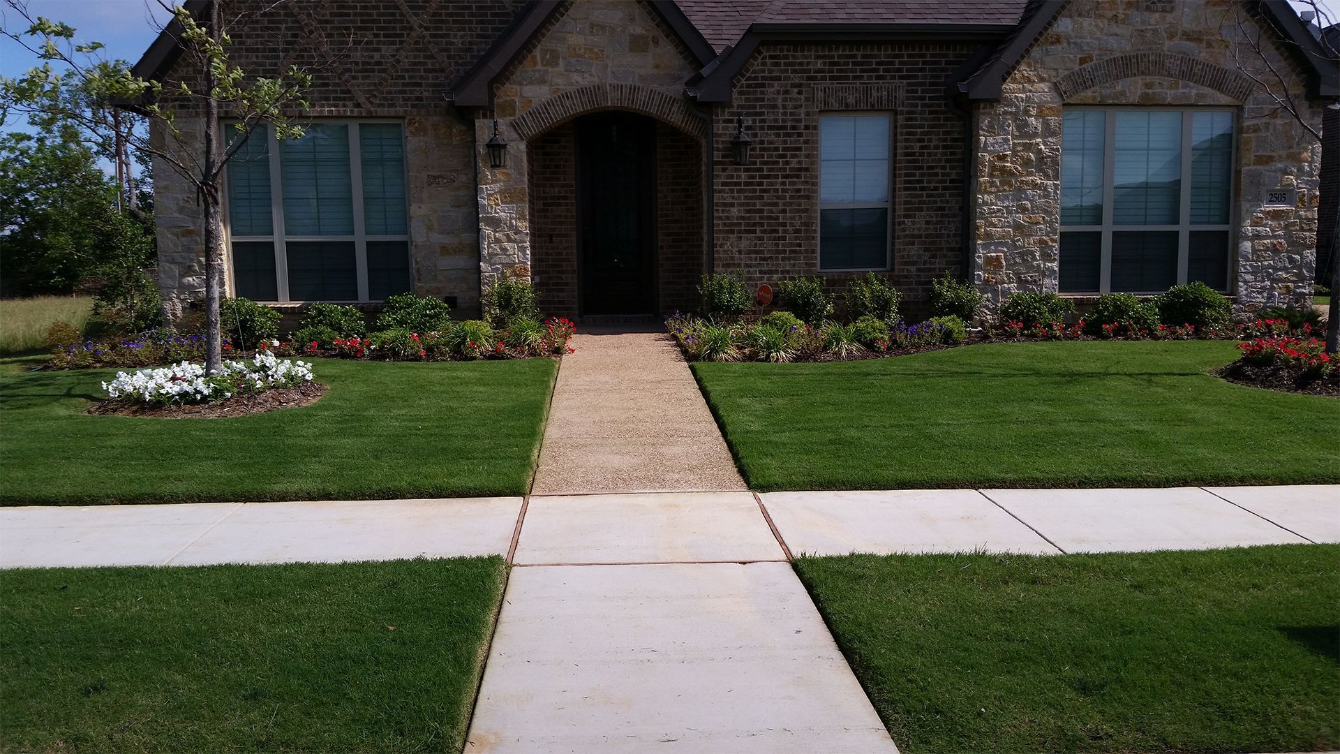 Brick house front with stone archway, central walkway, and manicured lawn and flower beds