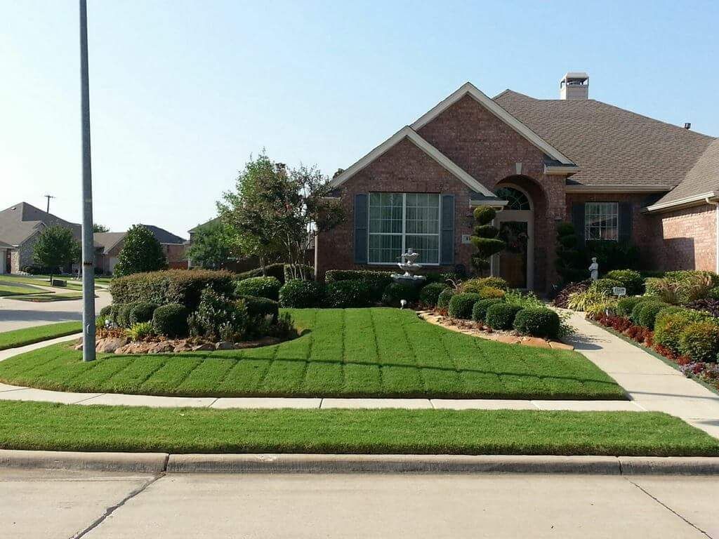 Brick house with manicured green lawn, shrubs, and a sidewalk in a quiet suburban neighborhood.