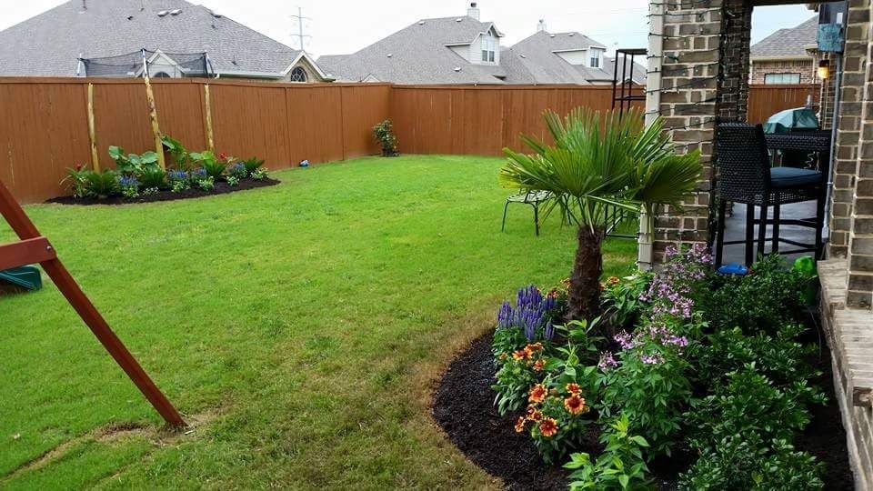 Backyard lawn with flower beds, a palm, wooden fence, and brick patio by a house