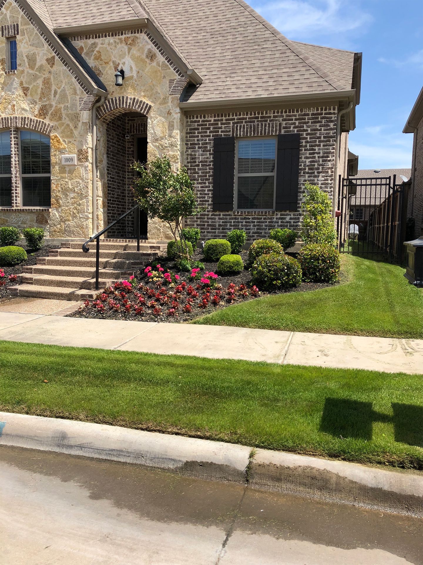 Stone house with front yard landscaping, red flowers, and a sidewalk by the curb.